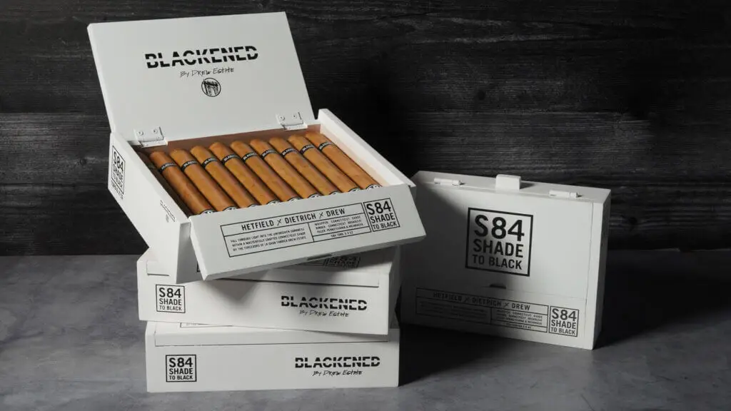 Display of multiple boxes of Blackened “S84 Shade to Black” by Drew Estate cigars, including an open box revealing the cigars inside, with bold black text on sleek white packaging.