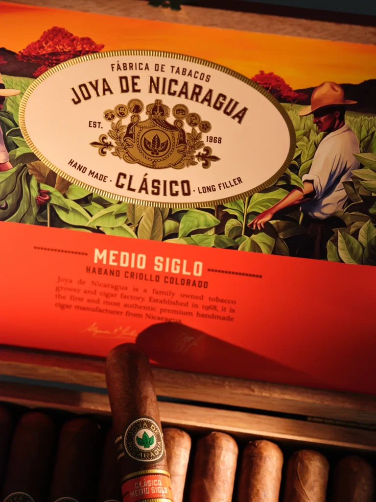 Box of Joya de Nicaragua Clásico cigars labeled “Medio Siglo Habano Criollo Colorado,” featuring an illustration of tobacco field workers and highlighting the brand’s heritage since 1968