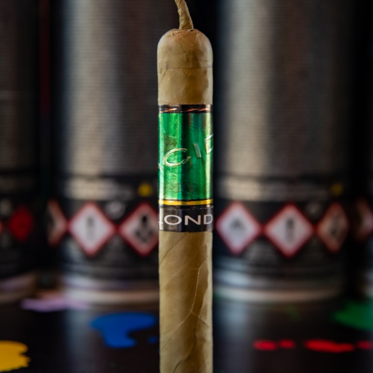 A Drew Estate Acid Green Cigar standing upright on a splatter-painted surface, featuring a light brown wrapper and a shiny metallic green band labeled 'ACID' above a black 'BLONDIE' band, with blurred spray paint cans in the background.
