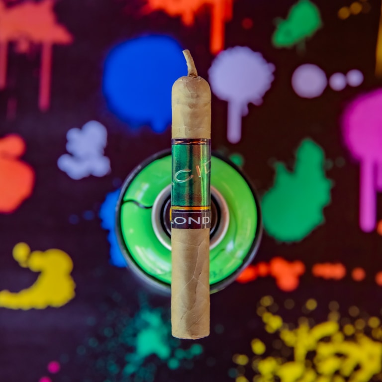 A Drew Estate Acid Green Cigar balanced upright on a green spray paint can, set against a black background splattered with vibrant colors like blue, pink, yellow, and purple.
