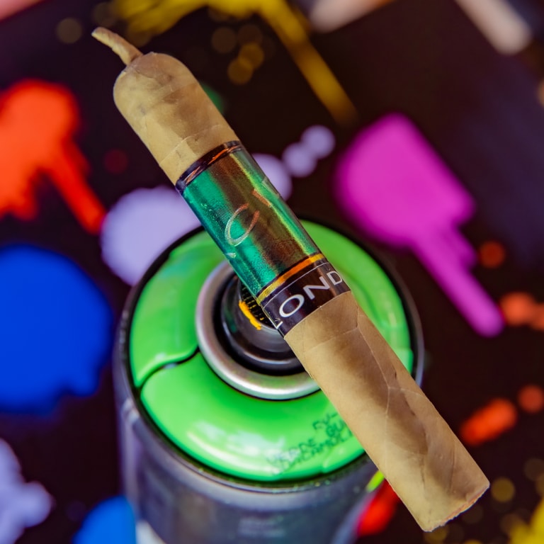 Drew Estate Acid Green Cigar resting horizontally on a green spray paint can, with a vivid backdrop of blue, red, and pink paint splatters.