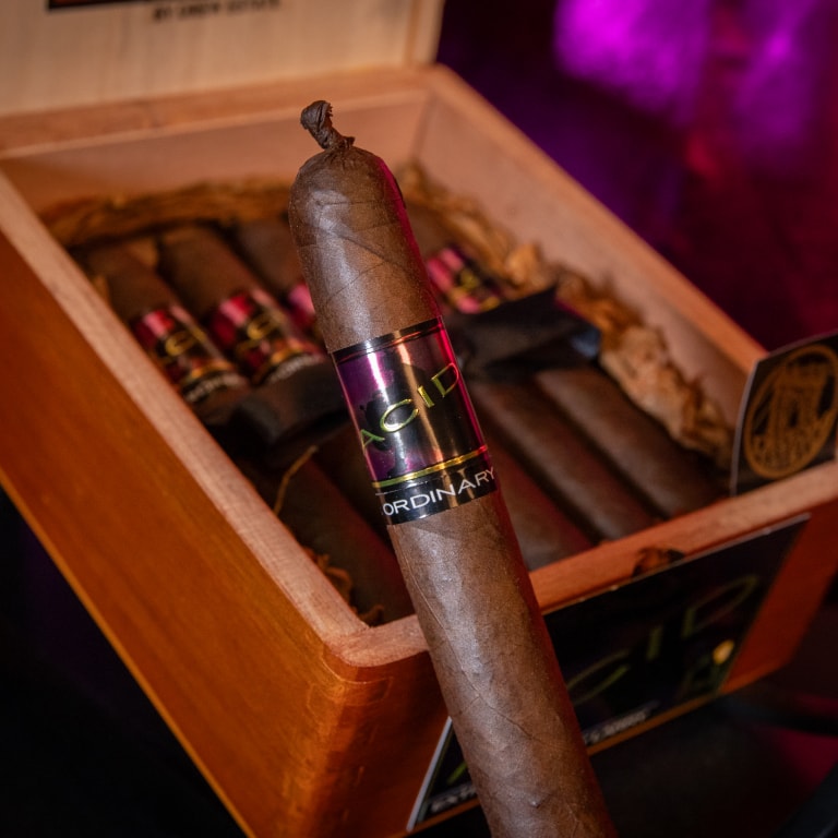 Close-up of a Drew Estate Acid Morado cigar labeled 'ACID' and 'ORDINARY,' resting in an open wooden box with other cigars in the background.