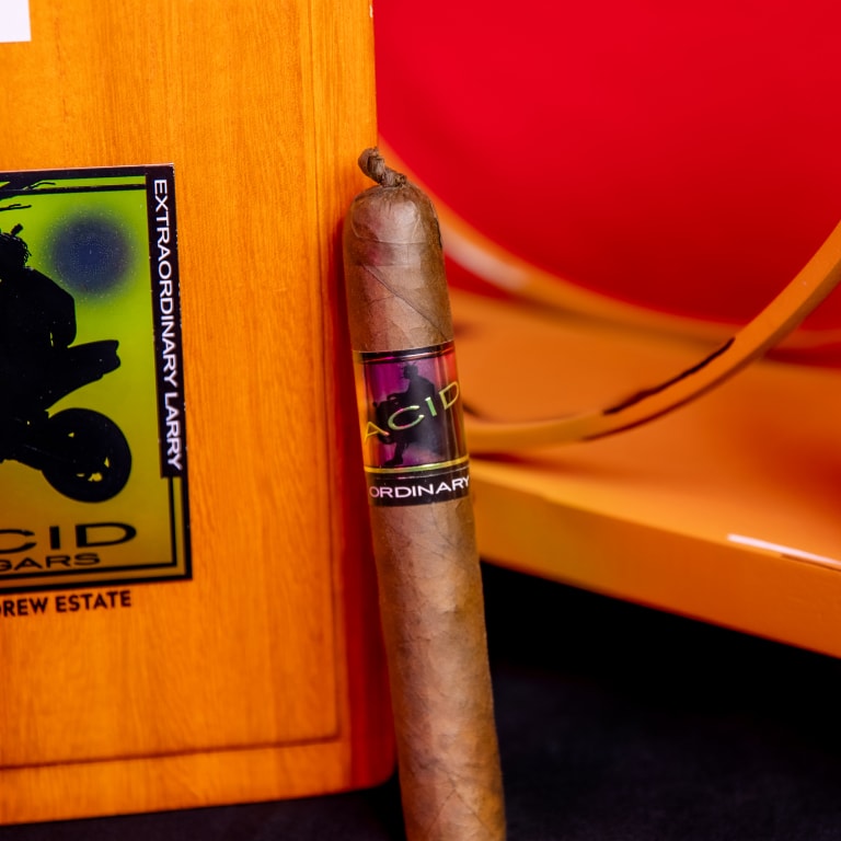 A Drew Estate Acid Morado cigar labeled 'ACID' and 'EXTRAORDINARY LARRY,' standing upright beside a matching wooden box with the same branding.