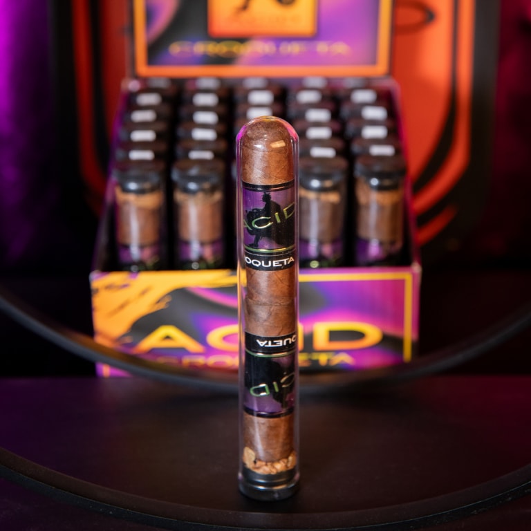 Close-up of a Drew Estate Acid Morado cigar with bands labeled 'ACID CROQUETA,' positioned in front of a matching display box.