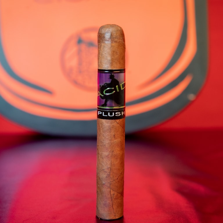 A single Drew Estate Acid Morado cigar standing upright on a red surface, featuring an 'ACID PLUSH' label with a silhouette design, set against an orange and black background.