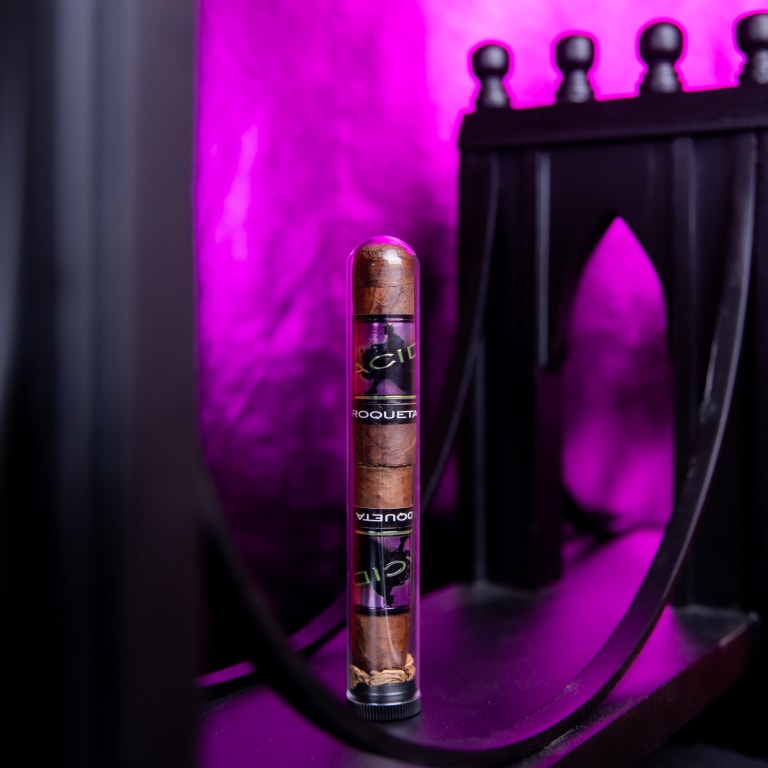A Drew Estate Acid Morado cigar encased in a clear tube with black and gold labels, set against a pointed arches and purple lighting.