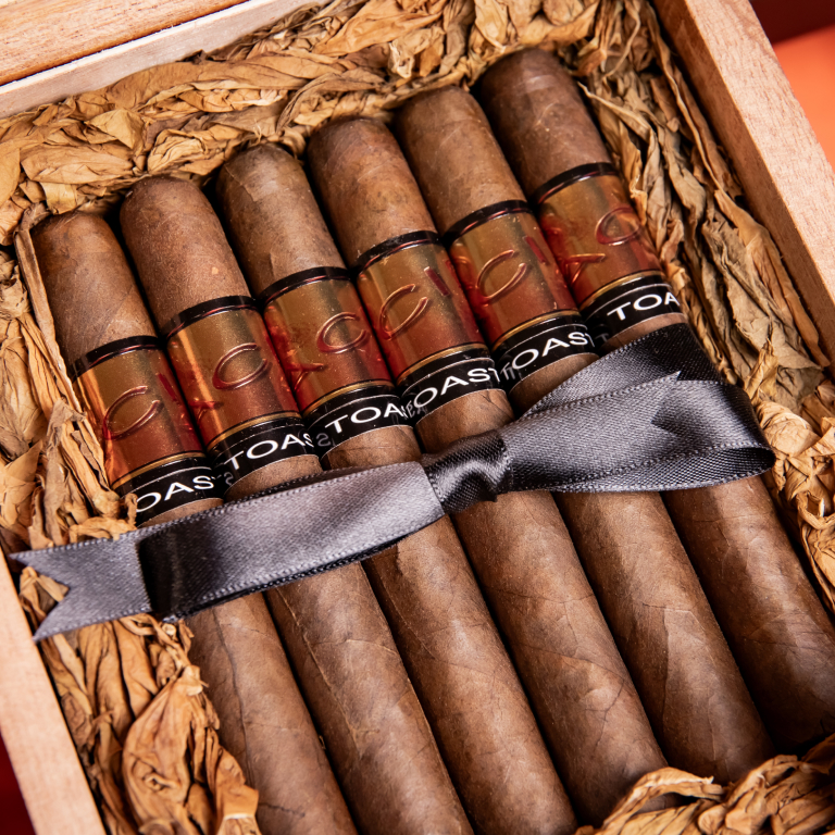 A wooden cigar box filled with six Drew Estate Acid Blue Cigars, specifically Toast Maduro cigars, nestled in shredded brown paper. Each cigar features a clear band with "TOAST" written in black, and a dark gray ribbon is tied across the center of the bundle.