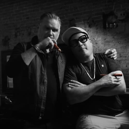 Two men standing together in a dimly lit setting, both holding cigars.