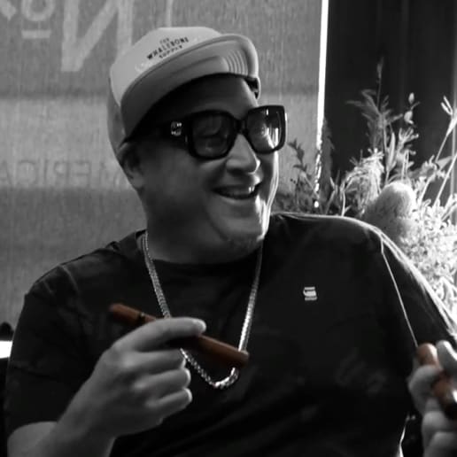 A smiling man in a hat and glasses holding a cigar