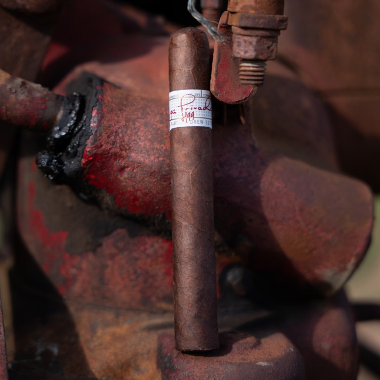 Close-up of a Liga Privada H99 cigar with a brown wrapper and a white band, resting against rustic machinery at the Connecticut Barn Smoker Event