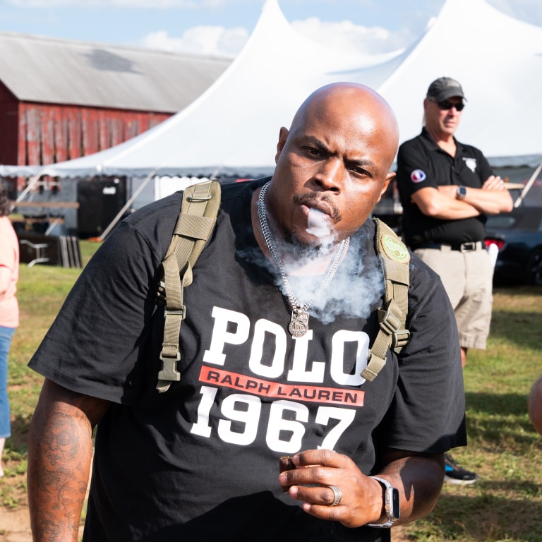 Man in a black Polo Ralph Lauren t-shirt surrounded by cigar smoke at the Connecticut Barn Smoker Event, with tents and a red barn structure in the background.