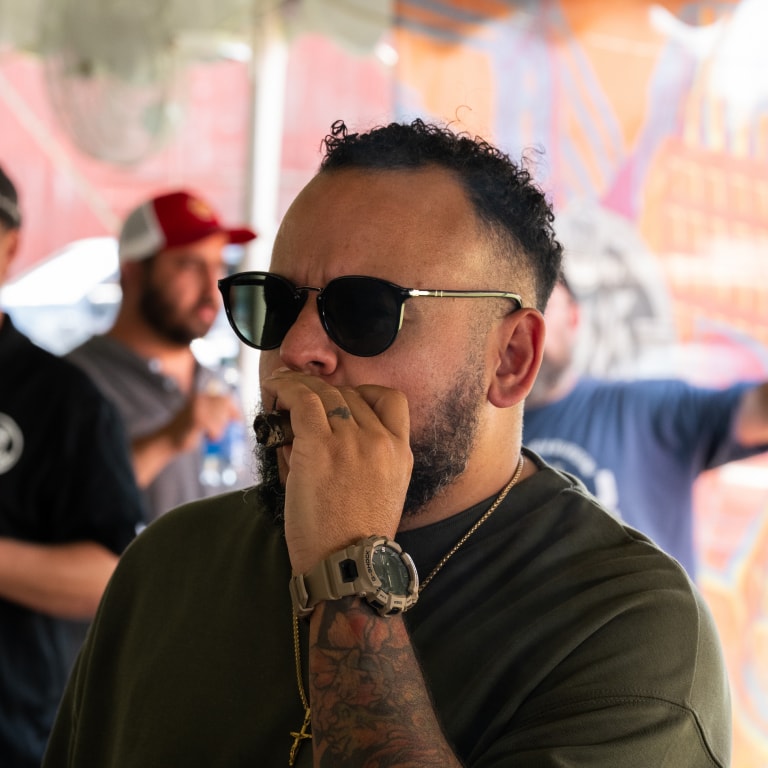 Tattooed attendee in sunglasses and a green shirt enjoys a cigar at the Connecticut Barn Smoker Event, with fellow participants in the background