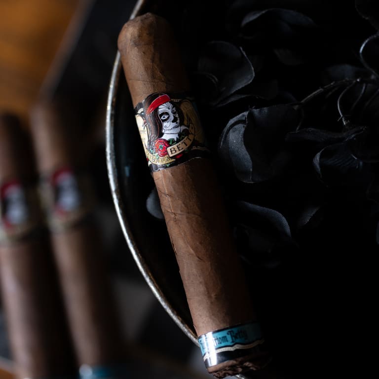 A Deadwood Fatbottom Betty cigar rests on a black bowl, set against a minimalist backdrop