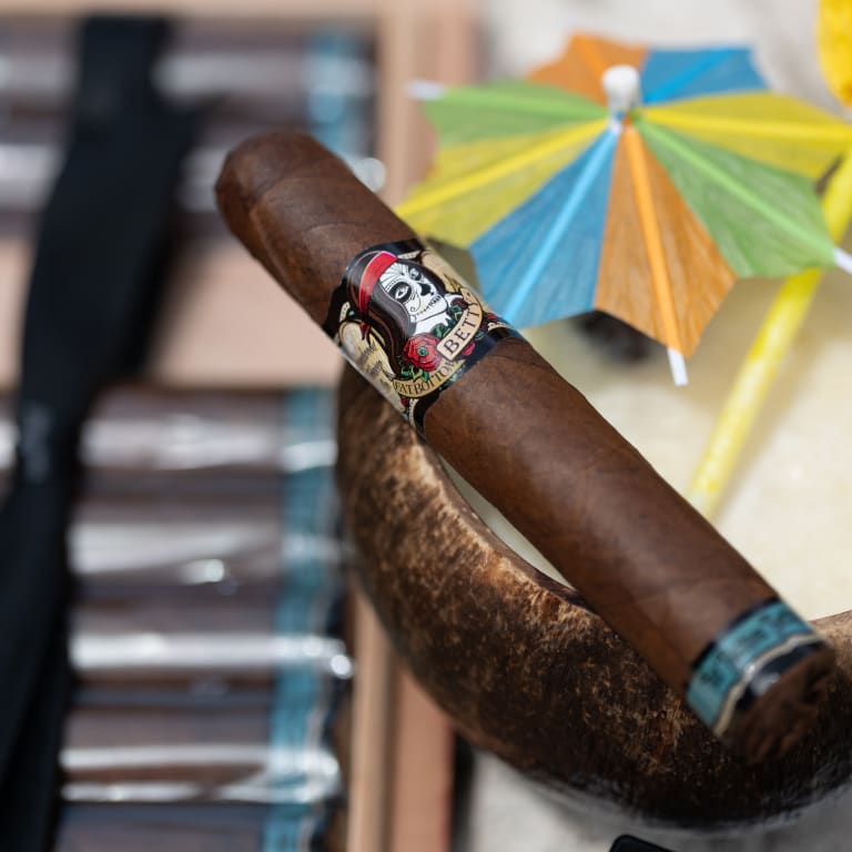 A Deadwood Fatbottom Betty cigar is placed on a table, next to a straw and umbrella