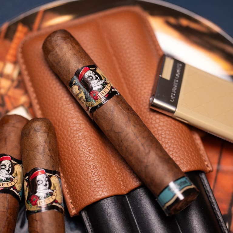 A Deadwood Fatbottom Betty cigar rests beside a leather cigar holder on a rustic wooden table