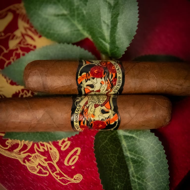Two Deadwood Sweet Jane cigars resting on a red table, adorned with leaves