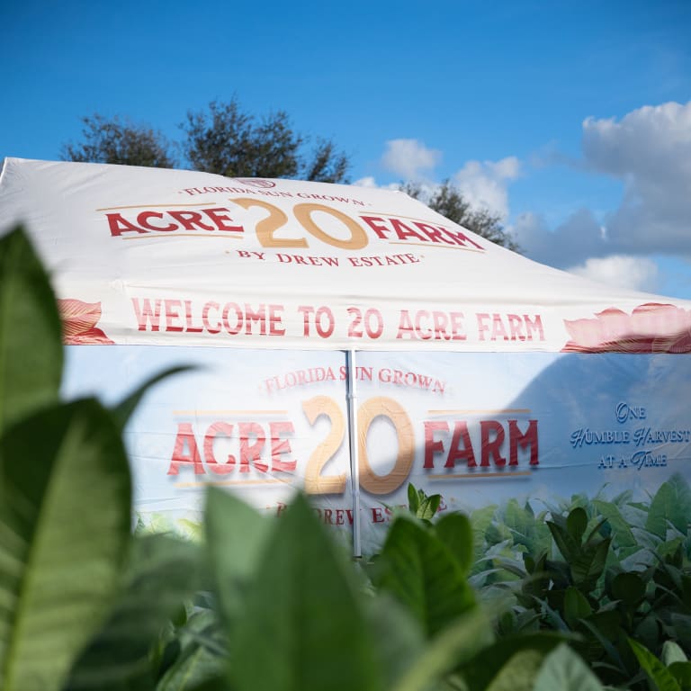 Tent featuring "Acre 20 Farm by Drew Estate" branding, surrounded by lush green plants under a bright blue sky