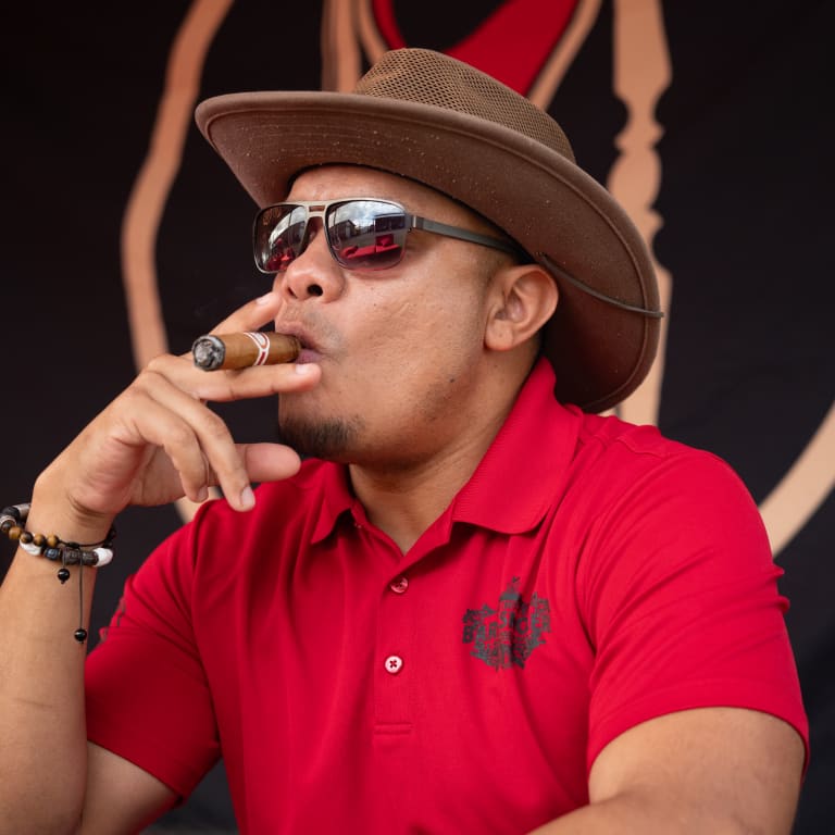 A man in a red polo shirt and cowboy hat enjoys a cigar at the Drew Estate Barn Smoker event