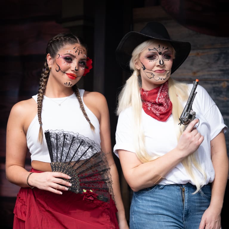 Two women in Día de los Muertos-inspired Western outfits, one with a lace fan, the other with a revolver prop