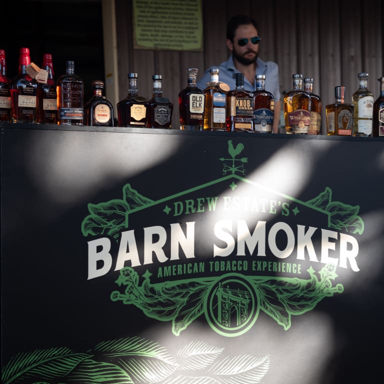 A bartender stands behind a display of premium whiskey bottles at Drew Estate’s Barn Smoker event