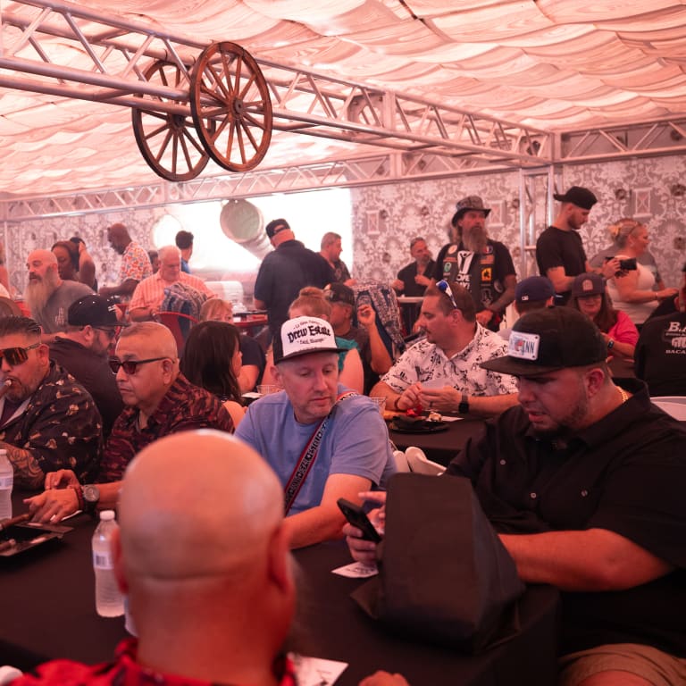 A lively crowd gathers under a decorated tent, enjoying conversations, cigars, and drinks at a social event