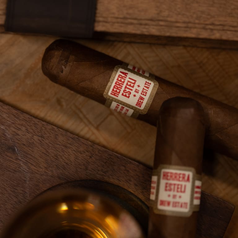 A Herrera Esteli cigar rests beside a glass of whiskey on a polished table