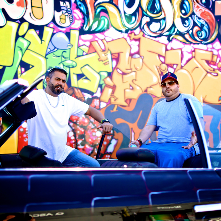 Willy Herrera and JD over an vintage car in front of a colorful graffiti wall