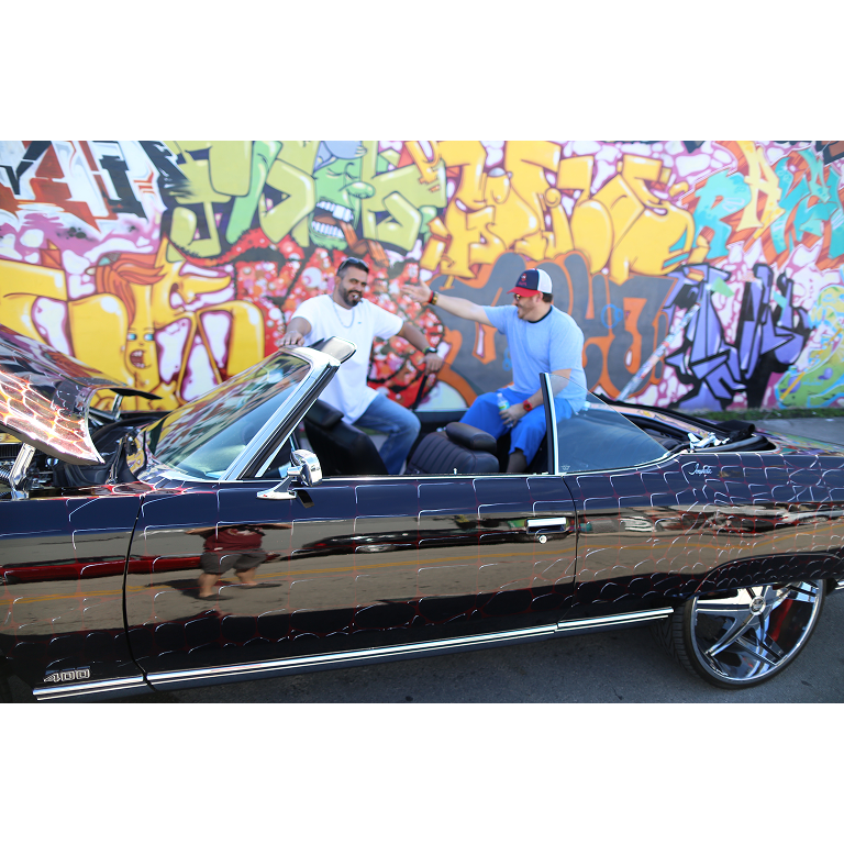 Willy Herrera and JD over an vintage car in front of a colorful graffiti wall