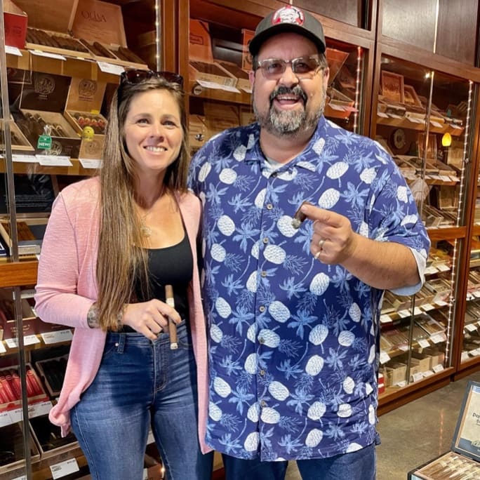 A man and woman pose together in front of a cigar store