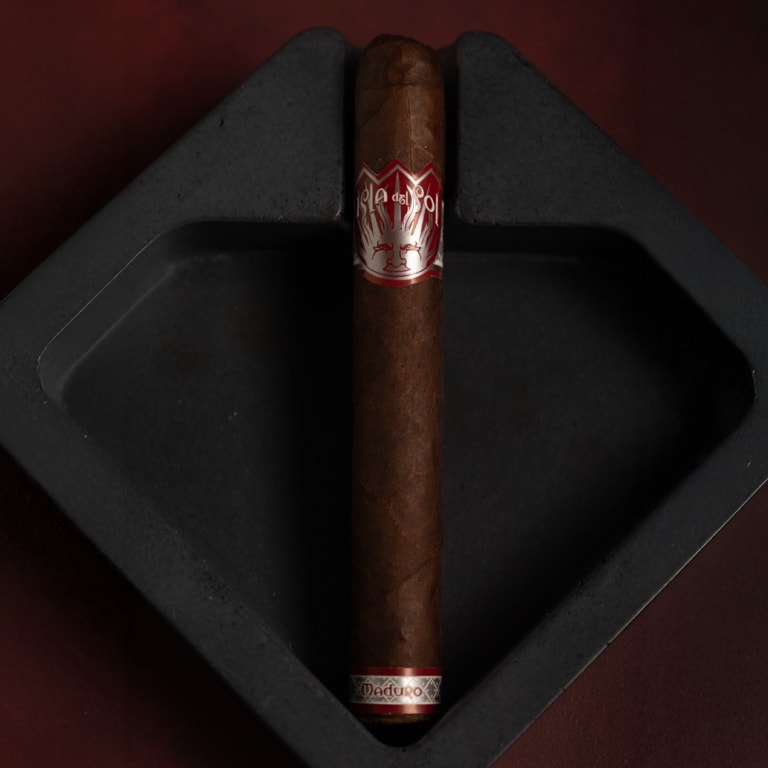 Isla del Sol Maduro cigar by Drew Estate resting in a sleek, black diamond-shaped ashtray. The cigar features two distinct bands—one red and white with the Isla del Sol branding, and a silver band labeled “Maduro”—highlighting its bold, rich character.