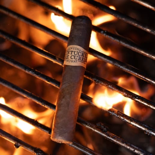 A Kentucky fire-cured cigar rests on a grill, surrounded by vibrant flames