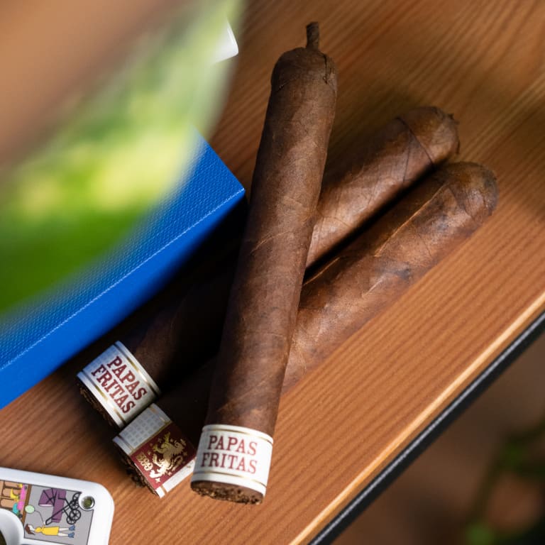 Three Liga Privada Papas Fritas cigars resting on a wooden table beside a vibrant green plant