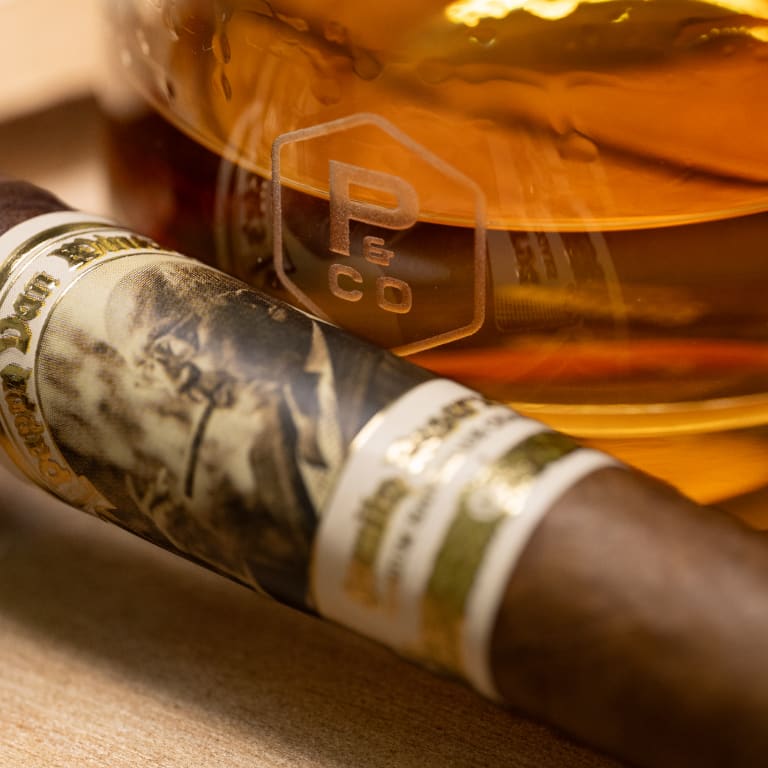 A Pappy Van Winkle barrel fermented cigar rests beside a bottle of whiskey on a polished table