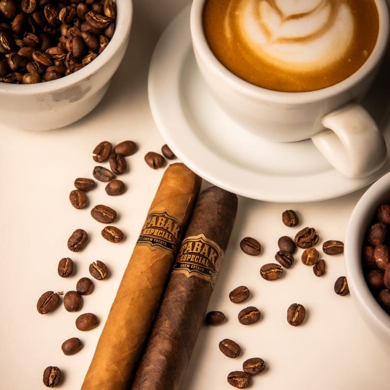 2 tabak especial cigars rest on a white surface beside a cup of coffee beans and cup of coffee