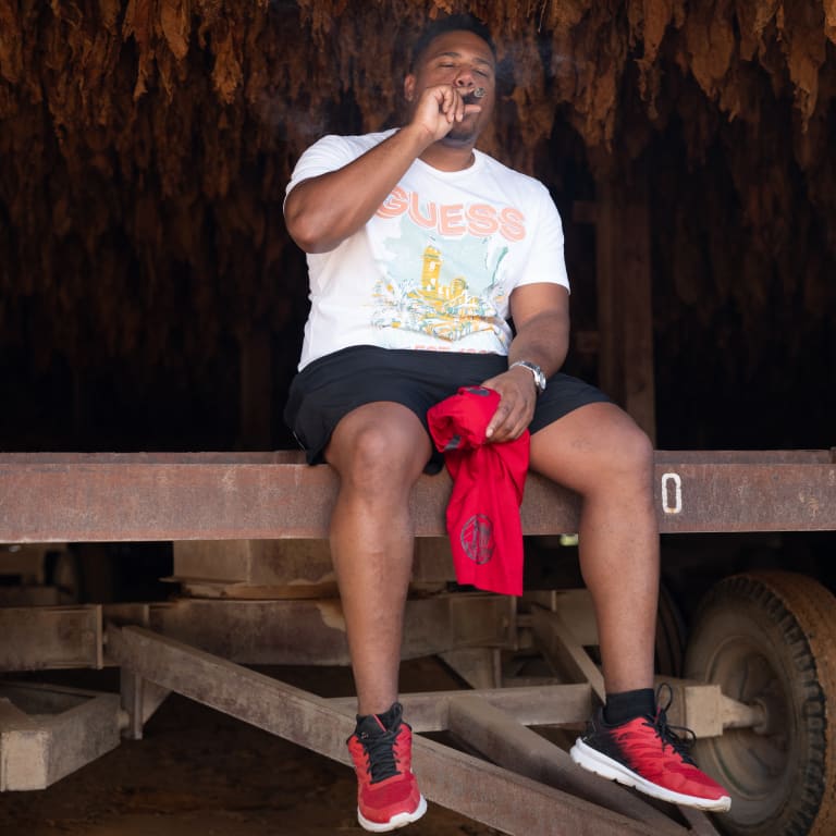 A man in a white tee and black pants sits on a bench, smoking a cigar with tobacco leaves in the background