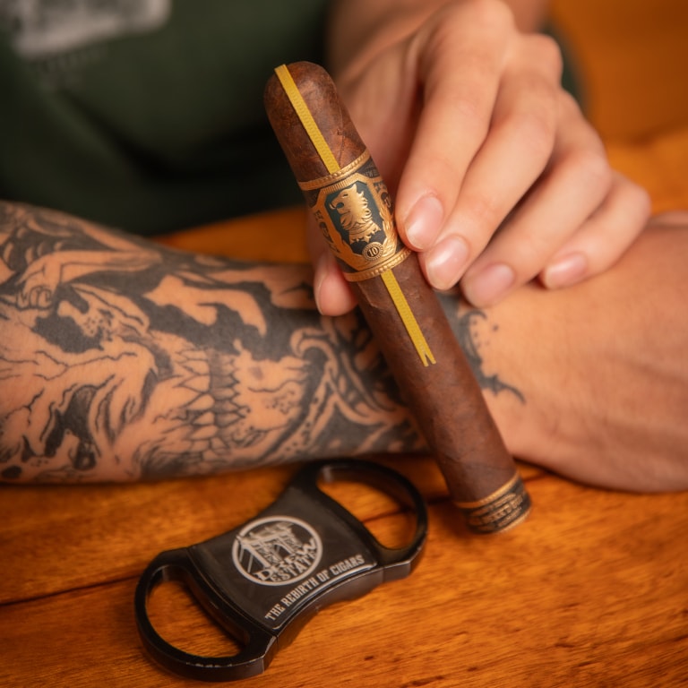 A tattooed man confidently holds a cigar in one hand and a cigar cutter in the other.