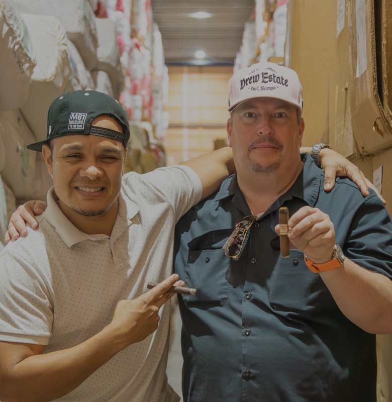 Two men holding cigars while standing in a warehouse surrounded by stacked boxes.