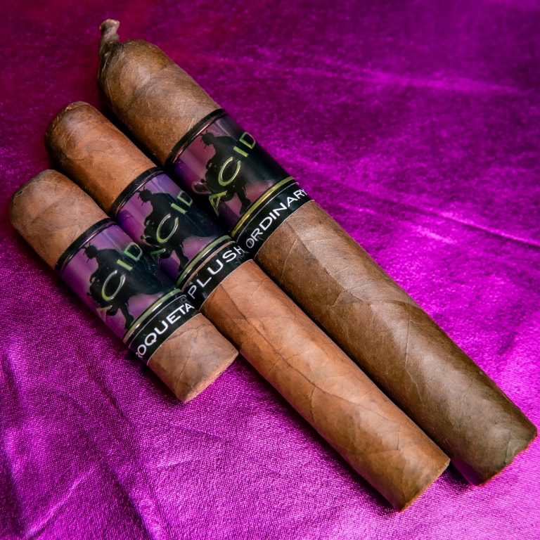 Set of three Drew Estate Acid Morado cigars displayed on a shiny purple background, emphasizing the bold branding and smooth wrappers.