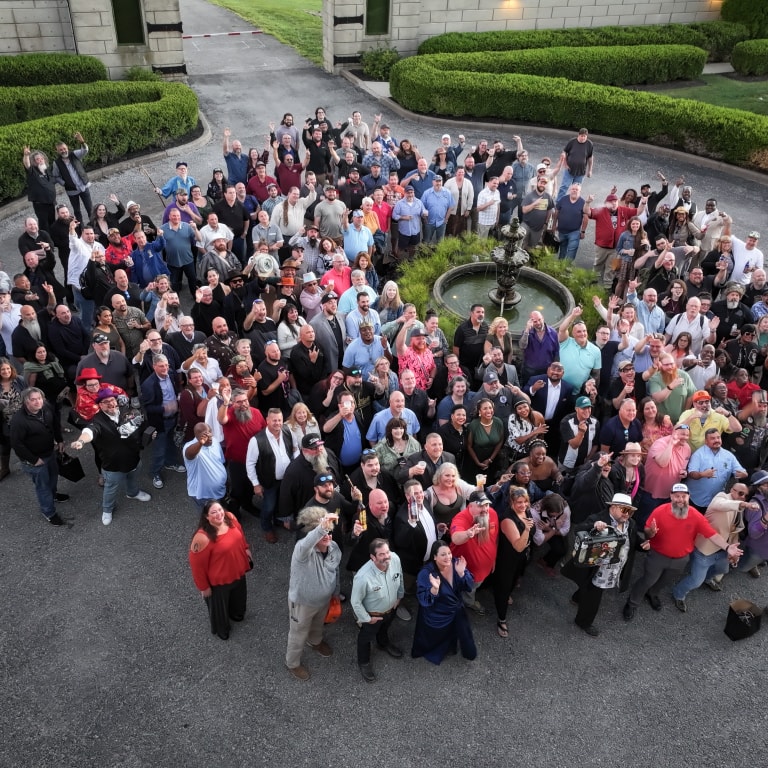 Outdoor group photo at the Kentucky Castle Family Reunion with people gathered near a fountain, waving joyfully in front of a castle-style building and hedges