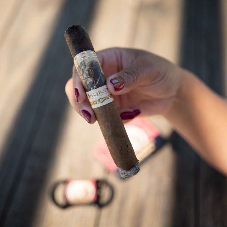 Hand holding a finely labeled Pappy Van Winkle cigar, red nails in focus, with a blurred wooden backdrop—an elegant detail from the Kentucky Castle Family Reunion.