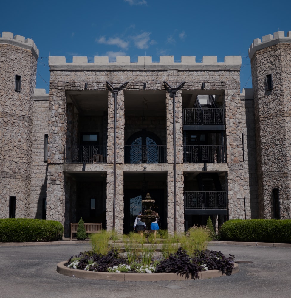 Front view of the Kentucky Castle with two cylindrical towers, a central arched entrance, and a fountain surrounded by a circular garden bed—setting for the Kentucky Castle Family Reunion.