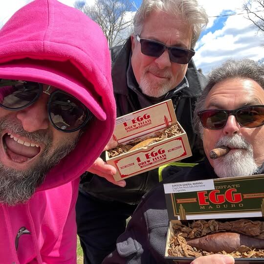 Three playful men holding two boxes of Drew Estate Egg cigars.