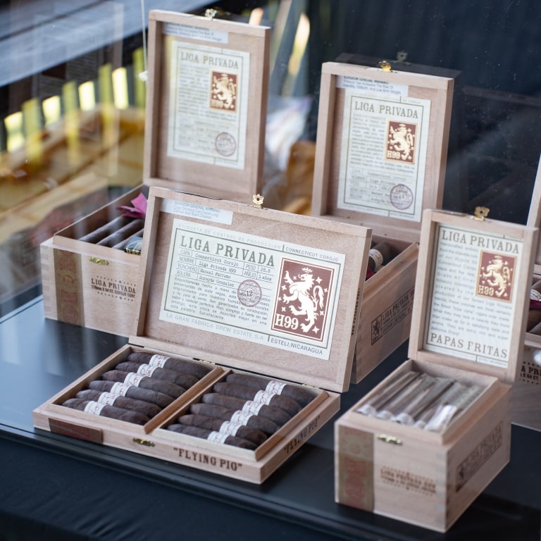 Open wooden cigar boxes from the Liga Privada collection displayed on a sleek black surface, featuring H99, Flying Pig, and Papas Fritas. Each box showcases intricate branding with lion emblems and detailed labels, capturing the essence of the Tour de Liga Experience