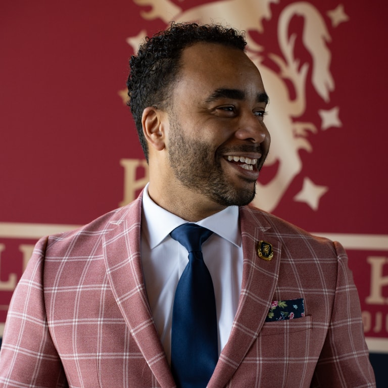 Happy man dressed in a stylish pink checkered suit jacket, white shirt, and navy tie, standing against a bold red backdrop featuring a lion emblem—capturing the sophisticated fashion and branding essence of the Tour de Liga Experience.