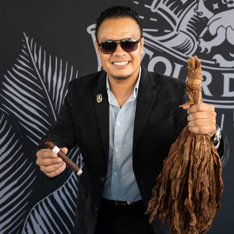 Person in a black blazer holding a Liga Privada cigar and a bundle of dried tobacco leaves, set against a black wall with the Tour de Liga logo.