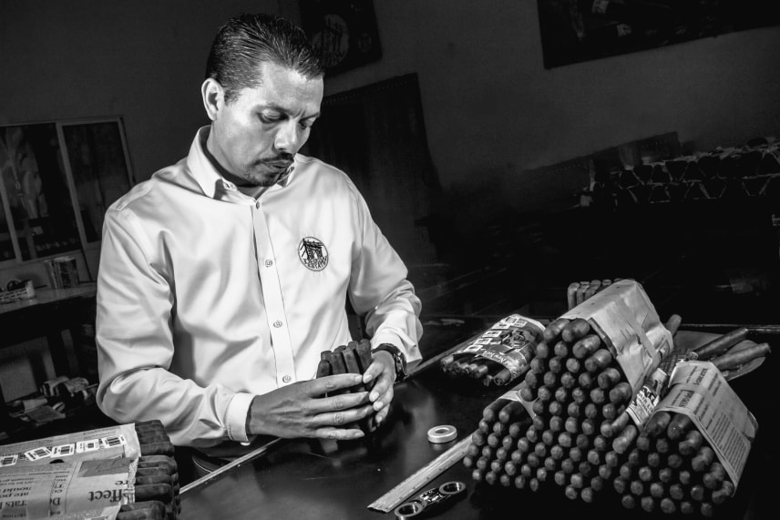 A black and white image of a man in a white shirt sitting at a table with a stack of cigars beside him.